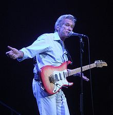 A man sings into a standing microphone, a guitar strapped around him and his arms spread outwards