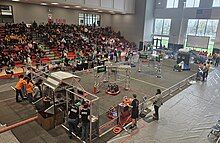 The Crescendo field during a match. Three red robots and two blue robots are visible, with one red robot sitting under its Stage. Another red robot is scoring at its Amp, and a blue robot is scoring at its Speaker