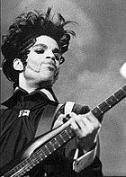 A young man with spiky hair and white face make-up holding an electric guitar; he is seen from a low viewpoint.