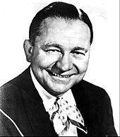 A dark-haired man wearing a dark jacket and a neckerchief