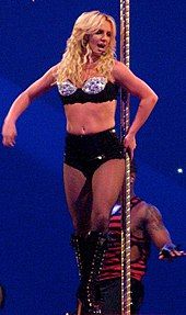 A female blond performer. She is leaning against a pole, grabbing it with her right hand. She is wearing a black sparkly bra, high-waisted shorts and laced-up high-heeled boots with fishnet stockings.