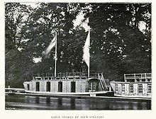 Two barges by the side of a river, one not fully in shot, in front of trees. The main barge has two large flags flying from the cabin roof, four large openings and one small one on the cabin's river side, and a window on the side of the cabin perpendicular to the river alongside a staircase to the roof; there are railings around the top of the roof. The other barge has five visible windows and roof railings.