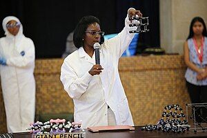 An African-American woman standing in a white lab coat holding a microphone to her mouth in her right hand, and a molecular model in her outstretched left hand