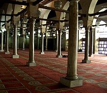 A large room filled with rows of cylindrical columns on top of square bases. The columns support arches which are pierced by square beams going the length of the room in both directions. Hanging from the beams are lamps, and the room's floor is covered with a red carpet with a repeated beige arched doorway shaped design on it. Exterior light enters from right of the room.