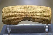 Rear view of a barrel-shaped clay cylinder resting on a stand. The cylinder is covered with lines of cuneiform text