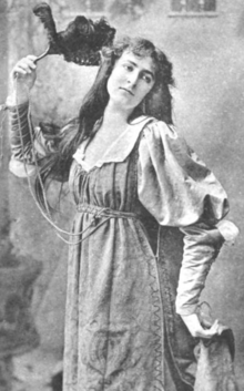 A young white woman, posed with a fan, holding one side of her skirt with her other hand; she is wearing a loose-fitting dress with round baggy upper sleeves and an open neckline; her hair is long and loose