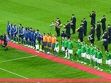 Several men standing on a grass football field. There are twenty five men standing in a straight line; eleven wearing blue on the left, three wearing orange tops and black shorts in the middle, and eleven wearing green on the right. In front of this line is a long narrow piece of red carpet. Behind the line are several middle aged or elderly men, wearing black and playing a variety of brass and percussion musical instruments.