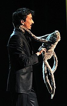 A man in a suit stands illuminated against a black background, holding a stuffed toy monkey.