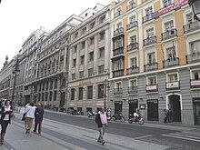 Photograph of 4 6-floor façades by a two-way street. 4 white people in autumn clothes walk on the pavement.