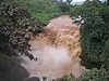 A brown waterfall cascades between rocks and heavy dark green vegetation.