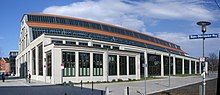 A long, large building with a barrel-vault roof rising above a facade with full-length plate-glass windows along the length of the building