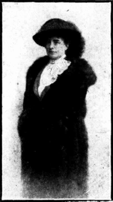 Black and white portrait of a woman standing, faced forward but to her left. She is wearing A white blouse with a high neck, a hat and a large fur coat which she has wrapped around her.