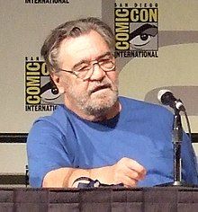 Head and upper torso view of a lightly-bearded, middle-aged man in glasses and a plain T-shirt, sitting behind a table with a microphone on it. Two small posters that read "Comic Con International" hang on the wall behind.