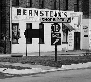 A black circle with the number 18 in white is mounted above a white-on-black arrow pointing to the upper right and a sign with the word east on a post at an intersection.