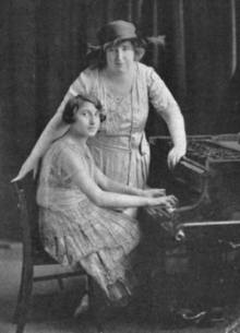 A young woman with dark bobbed hair is seated at a piano; an older woman wearing a brimmed hat is standing behind, one hand on chair, one hand on piano; both are wearing light-colored lacy gowns