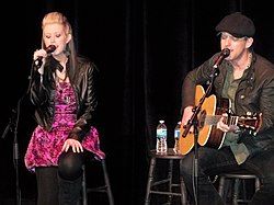 A woman with long blonde hair, wearing a purple dress, singing into a microphone, and a man wearing a cap singing and playing a guitar