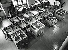 Looking down from above at a room at an intricate mechanical device which fills the room. In the background, a man sits at a desk next to a filing cabinet.[14]