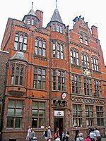 The front of a building with three storeys plus attics is seen from a slight angle. The ground floor is in stone; the upper storeys are in brick with stone dressings. On the front is a frieze containing heraldic shields, a coat of arms, and a variety of windows; at the top are two turrets and a shaped gable.