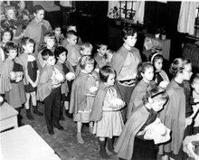 A group of small children and their teachers in a line, wearing brown capes and carrying food in their hands