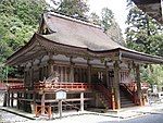 Three-quarter view of a proportionally tall wooden building with a veranda with red hand rail and a canopy covering the steps that lead to the central entrance. The roof appears to be a hip-and-gable roof.