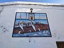 Photo from below of a white wall featuring 8 by 10 painted tiles. On the tiles, a boat or boats crewed by 11 white men. Some of the men are hauling two big tuna onto the boats. Brown nets cover the side of the boats into the water. Three white men on the net are partially into the water gesturing to the men on the boats. 6 tuna swim on the water near the nets. On the center top of the tiles there is a rectangle with a picture of a standing crowned woman holding a child and a scapular.