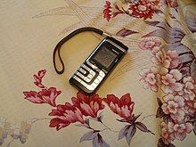 black Nokia 7260 cell/mobile phone with silver keys and a black wrist strap on a flowery table cloth