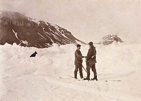 Two men shake hands in the midst of a snowfield, with a dog sitting nearby. Dark hills are shown in the background.