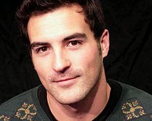 A photograph of a man with brown eyes, brown hair, and dimples looking at the viewer and wearing a green sweater over a black shirt