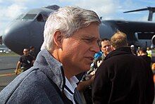 Image of Ambassador Swindells, right side profile, from the shoulders up. He's wearing a grey fleece jacket. Behind him, out of focus, are air force personnel and a grey C-141 Starlifter cargo plane.