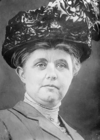 A black and white photograph of a woman wearing a large, decorative hat