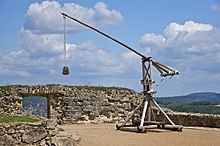 Photo of a reconstructed medieval trebuchet on a stone platform against a cloudy sky in the background