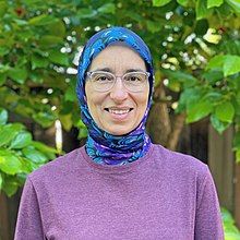 Photograph of a woman wearing a purple jumper, a blue headscarf, and glasses.