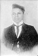 Head and torso of a young black man wearing a suit and tie with a watch chain hanging from a jacket button. He has a cap pushed high up on his forehead and tilted over his left ear.