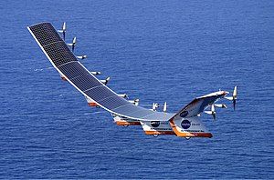 Helios Prototype in flight. The craft consists of a curved series of connected solar panels that have propellers behind them and fins on the bottom.