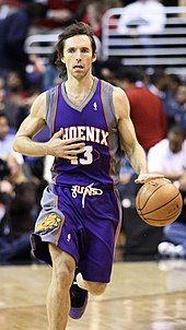 A white basketball player, wearing a purple jersey with a word "Phoenix" and the number "13" written in the front, dribbles the ball