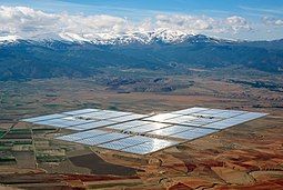 Concentrated solar power parabolic troughs in the distance arranged in rectangles shining on a flat plain with snowy mountains in the background