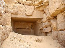 entrance of a stone lined corridor filed with desert sand amidst stone rubble