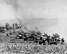 a row of trucks firing rockets. A cloud of dust is behind them.