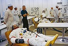 Several men in space suits recline on couches as others stand by.