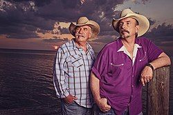 Two grey-haired men wearing cowboy hats, standing in front of the sea at sunset