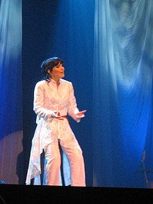 Turunen sings live onstage while seated on a stool. She is wearing a glittery white suit.