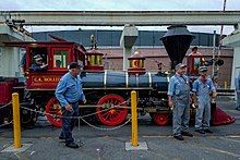 A red steam locomotive with a 4-4-0 wheel arrangement (four leading wheels, four driving wheels, and no trailing wheels) passing over a bridge