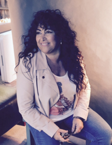 A woman with black curly hair and wearing a khaki jacket and blue jeans is sitting down and facing left