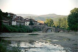 A river with a bridge spanning across it under a clear sky and greenery surrounding the water along with a few picturesque constructions.