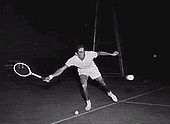 A man in white shorts and a white shirt swings a wooden racket with his right arm on a tennis court at night