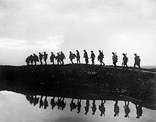 Soldiers walking across duckboards above a water-filled shell crater