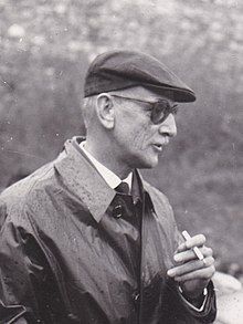 A black-and-white photo of a man in a flat cap and sunglasses holding a cigarette.