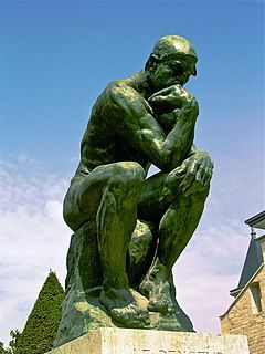 Photo depicting Rodin's statue The Thinker. The photo is taken obliquely from the front and from a frog's perspective. The sky in the background is blue.