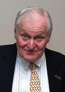 A man with white hair, seen from the chest up, looks directly into the camera with a surprised or excited facial expression. He is wearing a white collared shirt with thin multi-color vertical stripes, a black jacket with a "Brooklyn" pin, and a light-yellow tie with a repeating pattern of cherries.