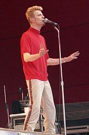 A middle-aged man with spiked hair and a red shirt singing next to a microphone